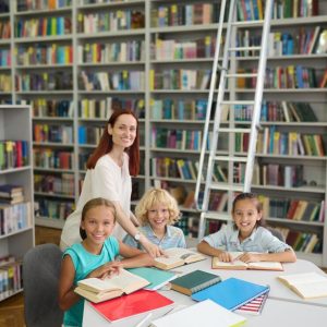 reading-library-young-smiling-woman-standing-near-table-touching-open-book-with-hand-joyful-boy-girls-school-age-looking-camera_259150-58175